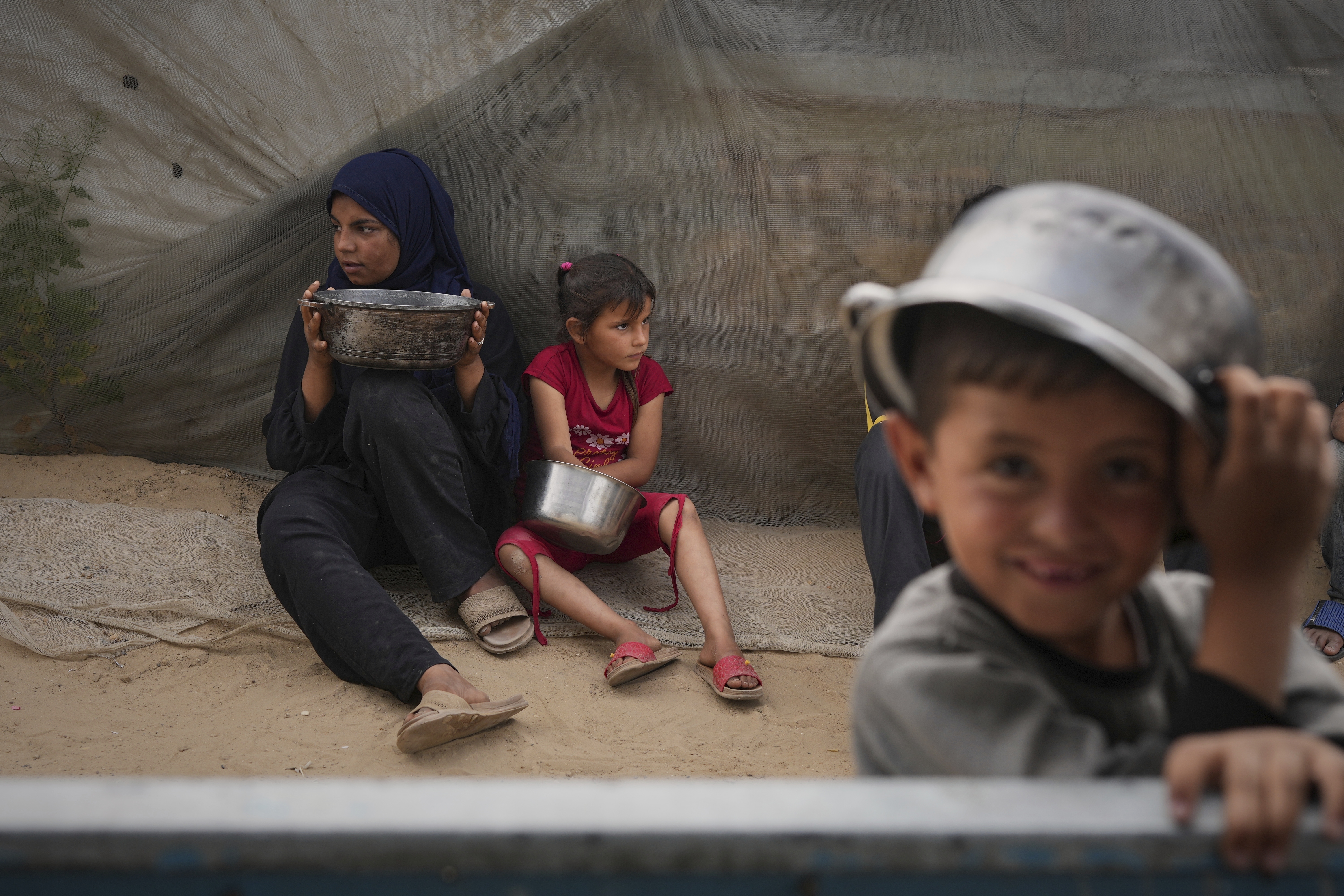 Mira Abu Shaar, 5, right, and her older sister, Raghad, 15, hold pots next to their family tent, as they wait for food to be prepared, in Muwasi, on the outskirts of Khan Younis in the southern Gaza Strip, Thursday, April 24, 2025. (AP Photo/Abdel Kareem Hana)