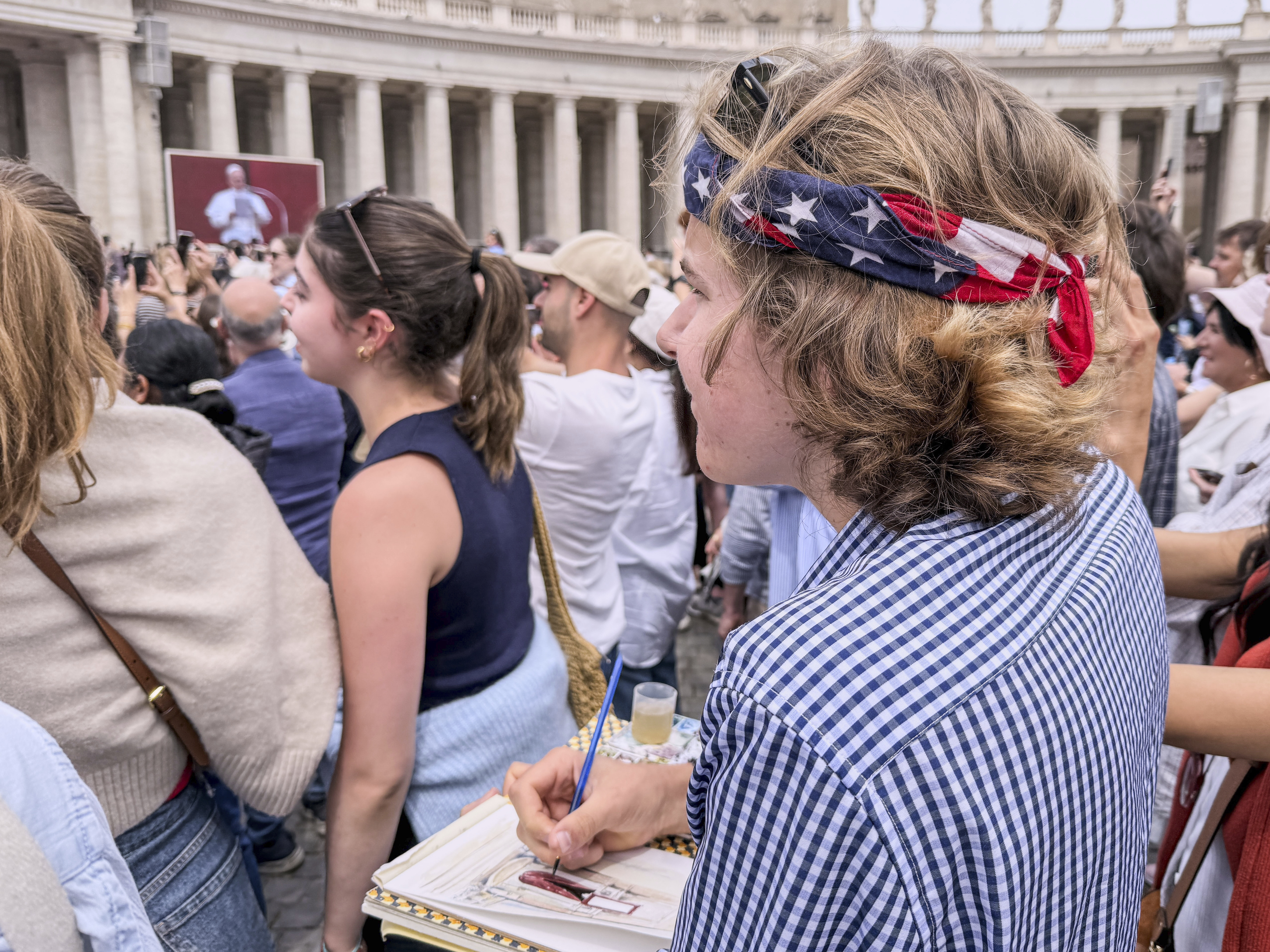 Notre Dame's architecture college student Wesley Buonerba, wearing a us flag bandanna, does a watercolor sketch of the loggia of St. Peter's Basilica during Pope Leo XIV first Sunday blessing of faithful gathered in the square for the traditional blessing, Sunday, May 11, 2025. (AP Photo/Giovanna Dell'Orto)