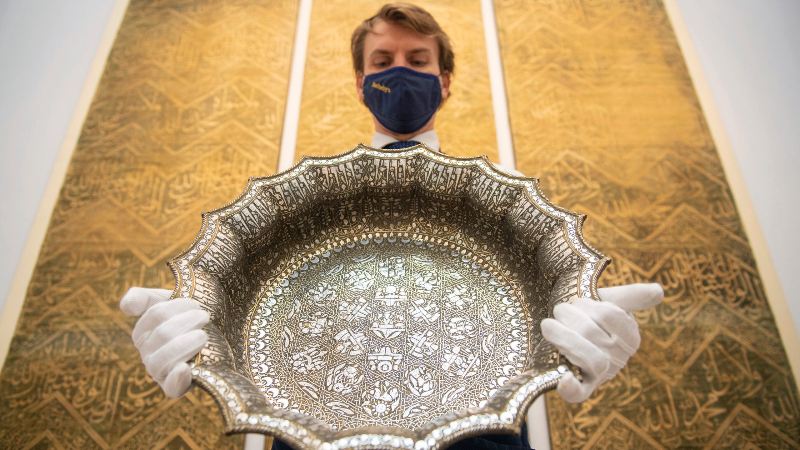 FILE - An employee at Sotheby's auction house holds a twelfth-century silver inlaid scalloped basin with astrological designs, estimated to fetch 1,000,000 - 1,500,000 sterling pounds, as part of the auction house's forthcoming Arts of the Islamic World and India sale on March 31, in London, March 29, 2021. (Dominic Lipinski/PA via AP)