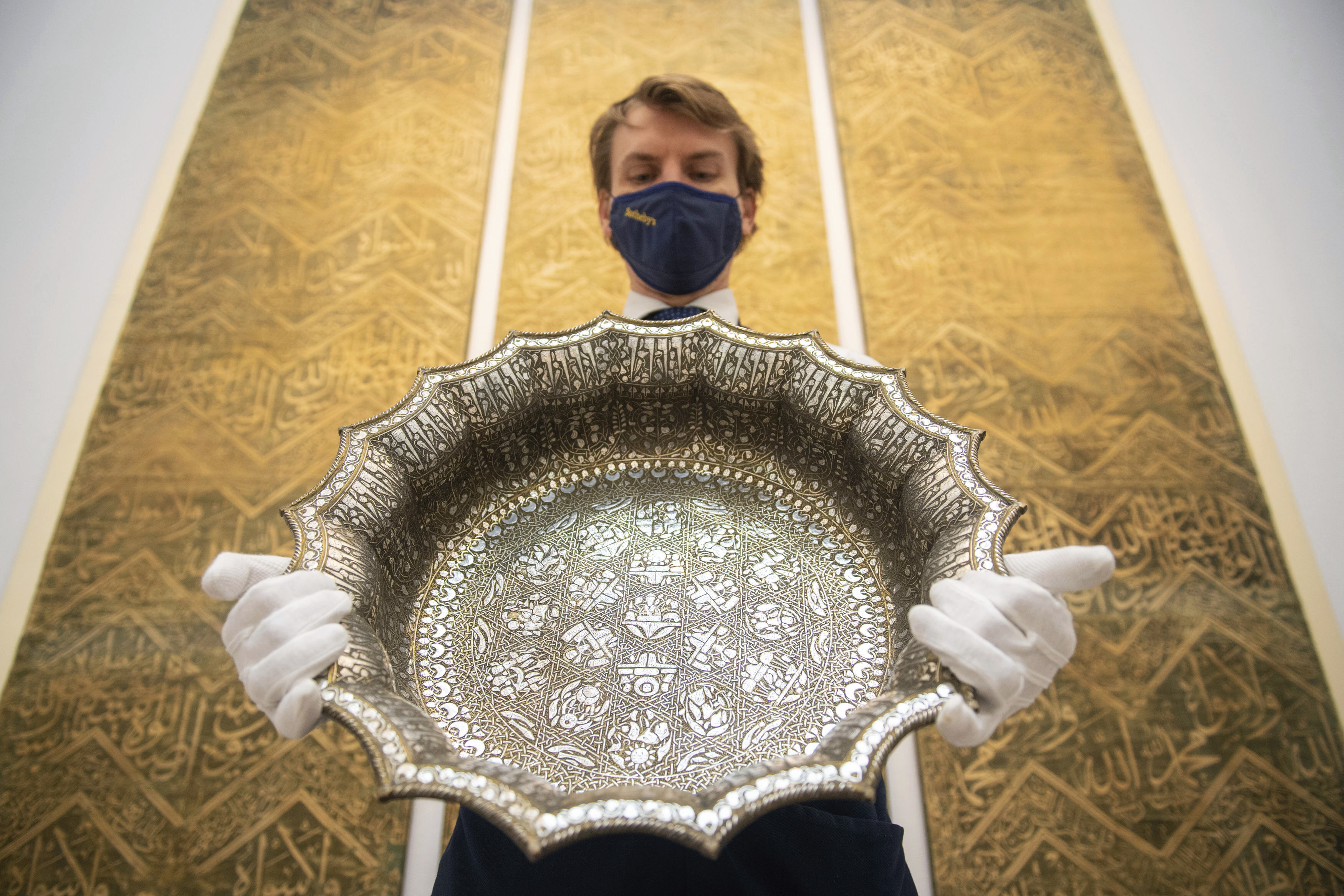 FILE - An employee at Sotheby's auction house holds a twelfth-century silver inlaid scalloped basin with astrological designs, estimated to fetch 1,000,000 - 1,500,000 sterling pounds, as part of the auction house's forthcoming Arts of the Islamic World and India sale on March 31, in London, March 29, 2021. (Dominic Lipinski/PA via AP)