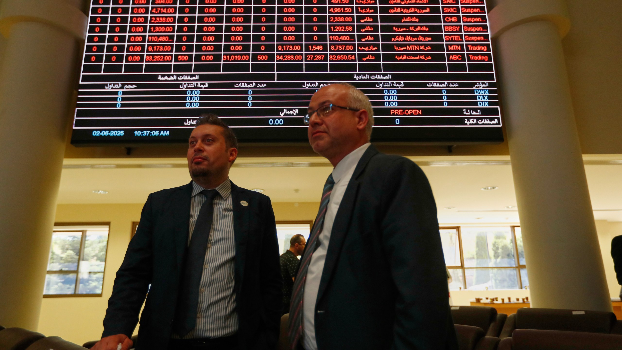 Two men stand under an electronic board showing stock prices at the Damascus Securities Exchange in Damascus, Syria, Monday, June 2, 2025. (AP Photo/Omar Sanadiki)
