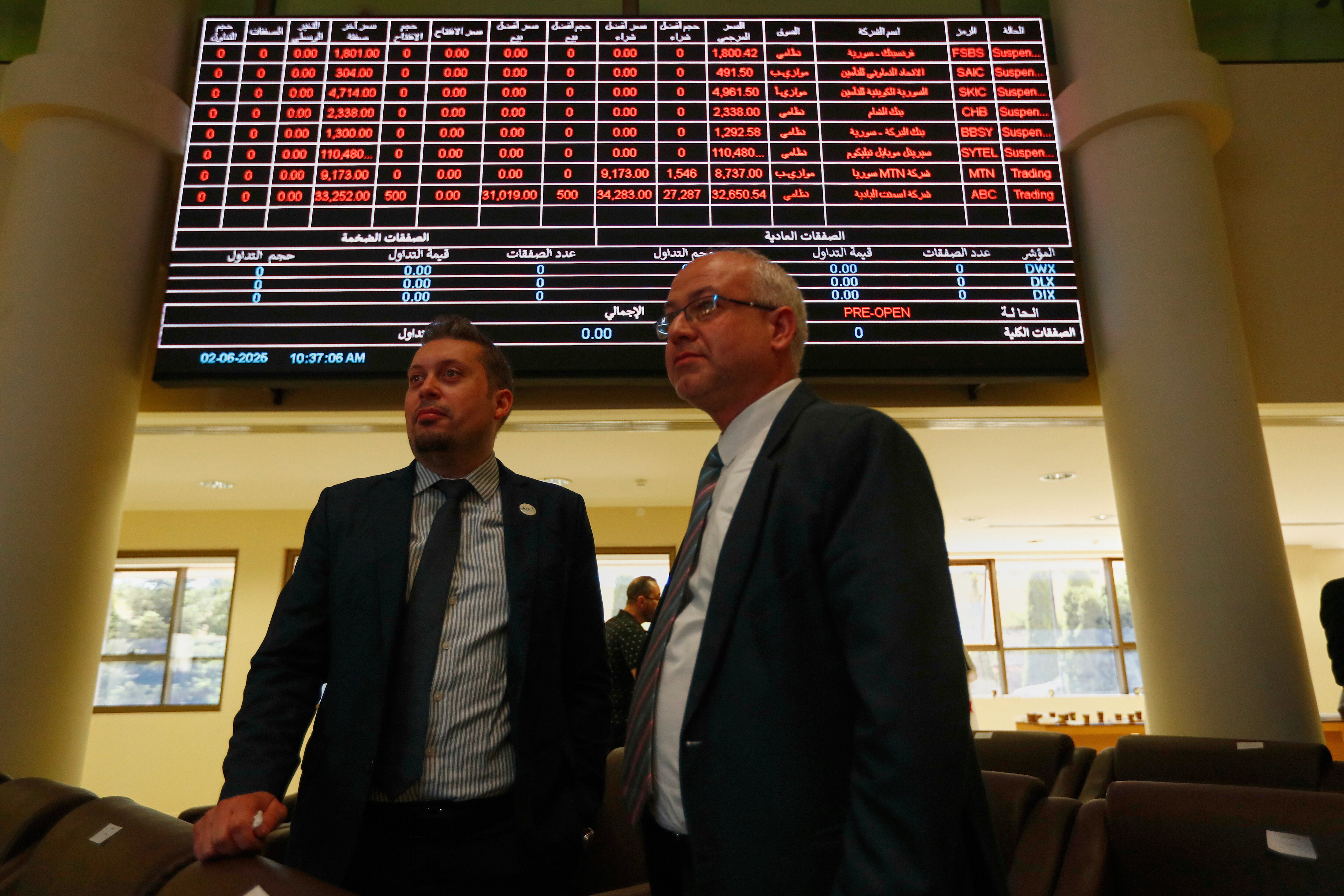 Two men stand under an electronic board showing stock prices at the Damascus Securities Exchange in Damascus, Syria, Monday, June 2, 2025. (AP Photo/Omar Sanadiki)