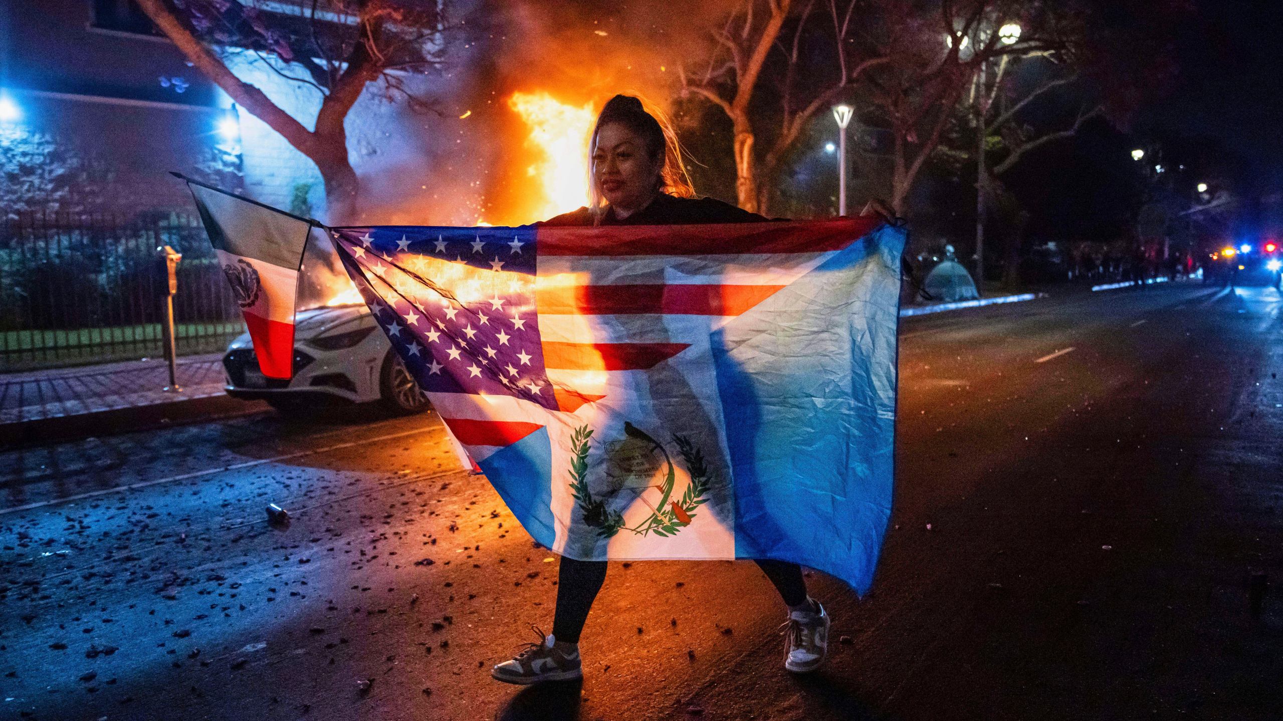 A person displaying multiple flags walks past a burning car during protests over the Trump administration's immigration raids in Los Angeles, Monday, June 9, 2025. (AP Photo/Ethan Swope)