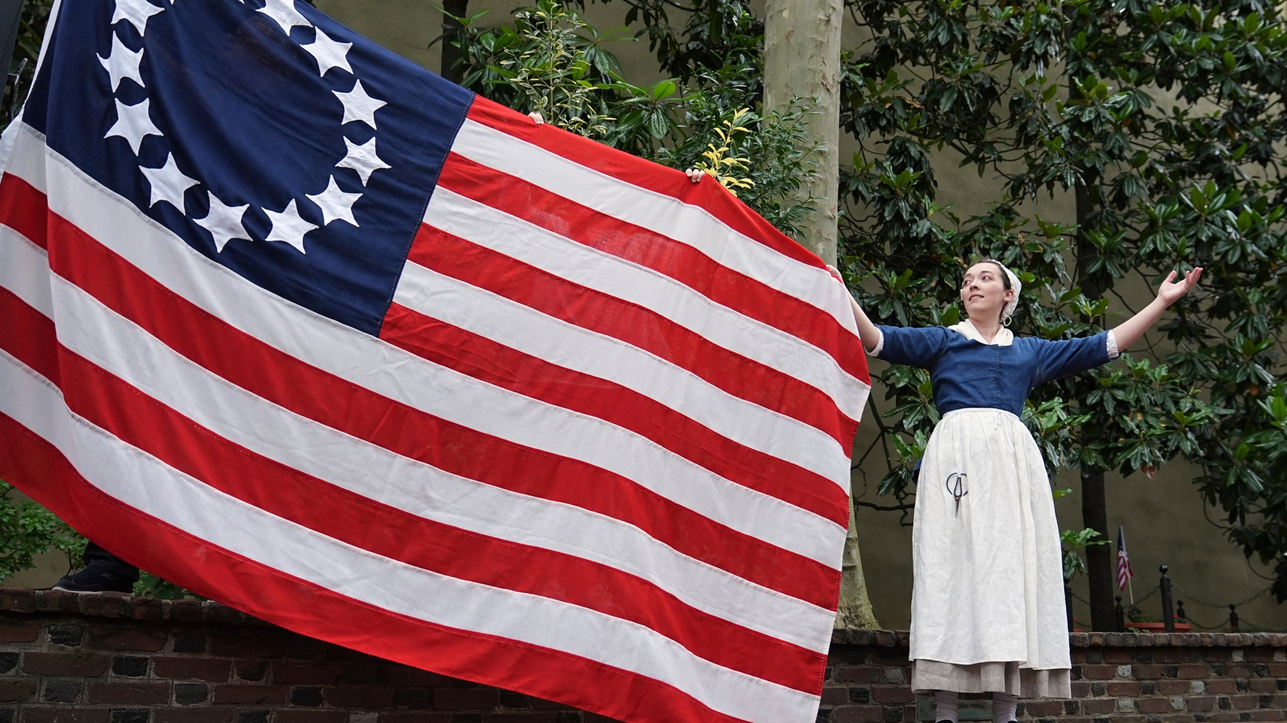 Madeline Burk, portraying Betsy Ross, holds a flag as it is hoisted up at the Betsy Ross House in Philadelphia, Monday, June 9, 2025. (AP Photo/Matt Rourke)