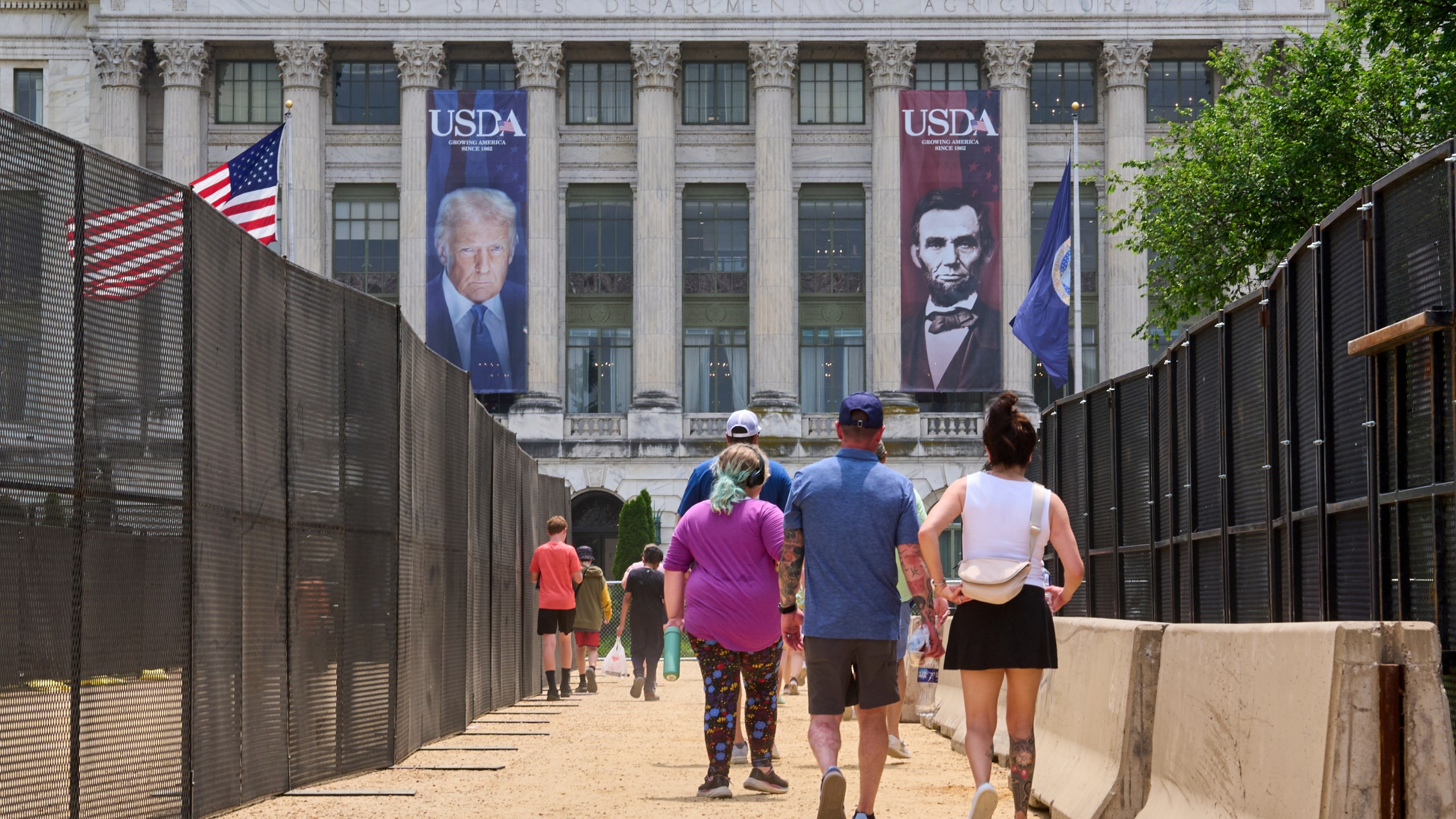 People walk in between security fencing and concrete barricades toward a large photograph of President Donald Trump and President Abraham Lincoln, draped on the USDA building, Friday, June 13, 2025, on the National Mall in Washington. (AP Photo/Jacquelyn Martin)