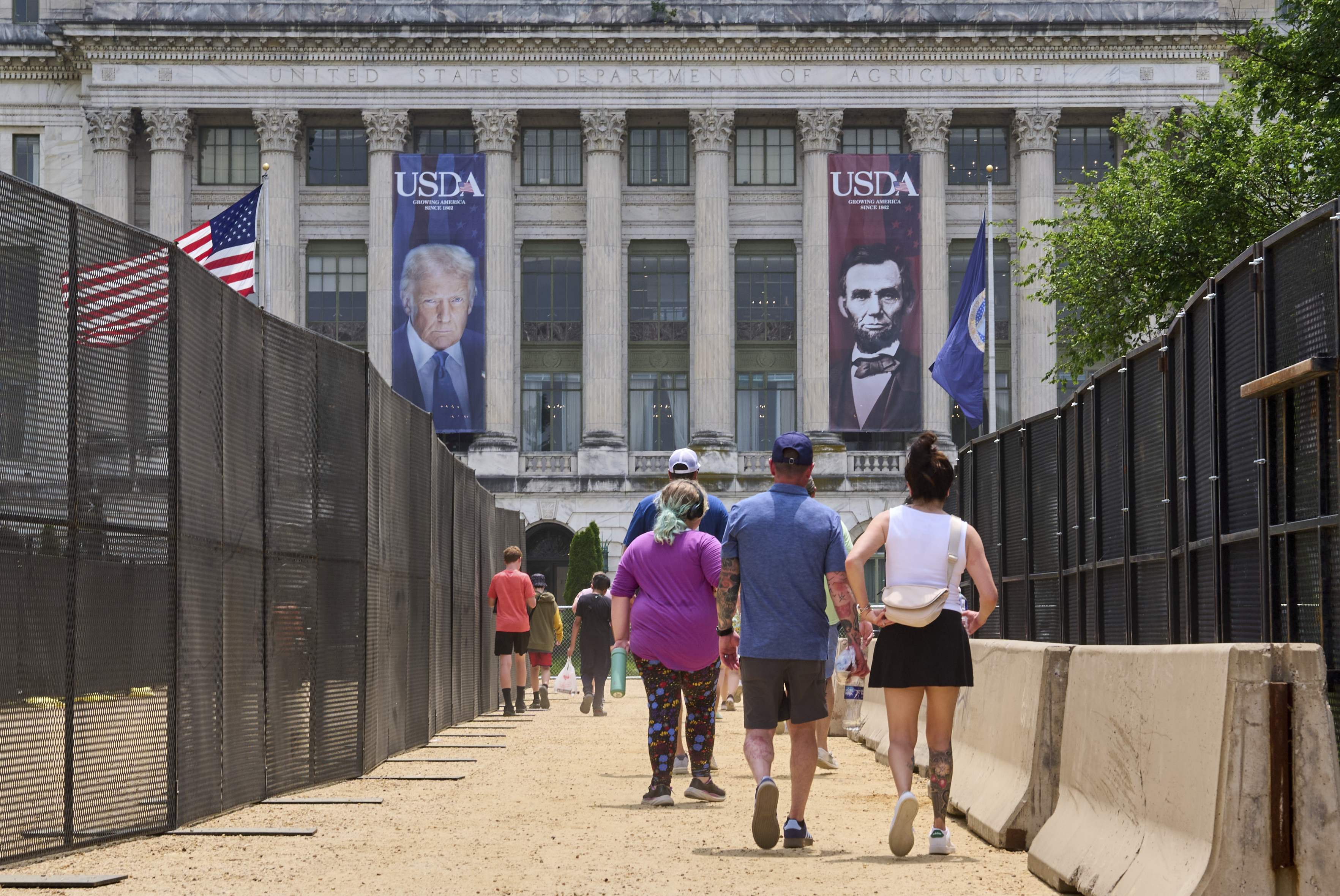 People walk in between security fencing and concrete barricades toward a large photograph of President Donald Trump and President Abraham Lincoln, draped on the USDA building, Friday, June 13, 2025, on the National Mall in Washington. (AP Photo/Jacquelyn Martin)