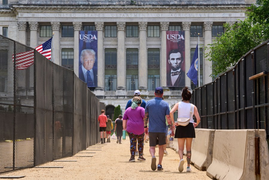 People walk in between security fencing and concrete barricades toward a large photograph of President Donald Trump and President Abraham Lincoln, draped on the USDA building, Friday, June 13, 2025, on the National Mall in Washington. (AP Photo/Jacquelyn Martin)