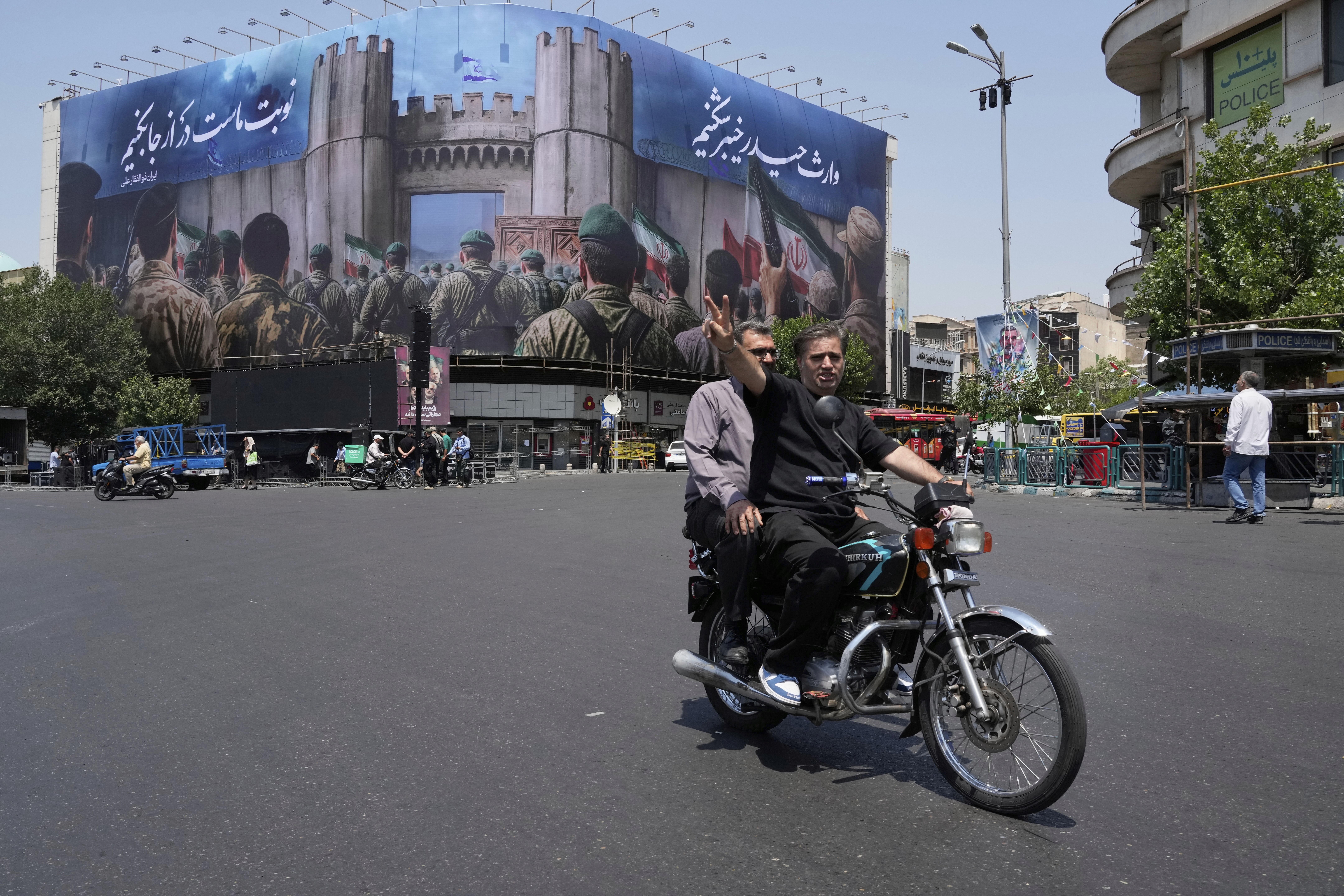 A man flashes a victory sign while riding his motorbike past an anti-Israeli banner depicting Iranian soldiers heading to attack Israeli territory, at Islamic Revolution Square in Tehran, Iran, Saturday, June 14, 2025. (AP Photo/Vahid Salemi)