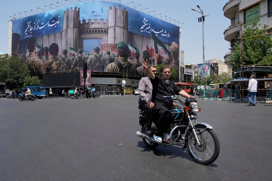 A man flashes a victory sign while riding his motorbike past an anti-Israeli banner depicting Iranian soldiers heading to attack Israeli territory, at Islamic Revolution Square in Tehran, Iran, Saturday, June 14, 2025. (AP Photo/Vahid Salemi)