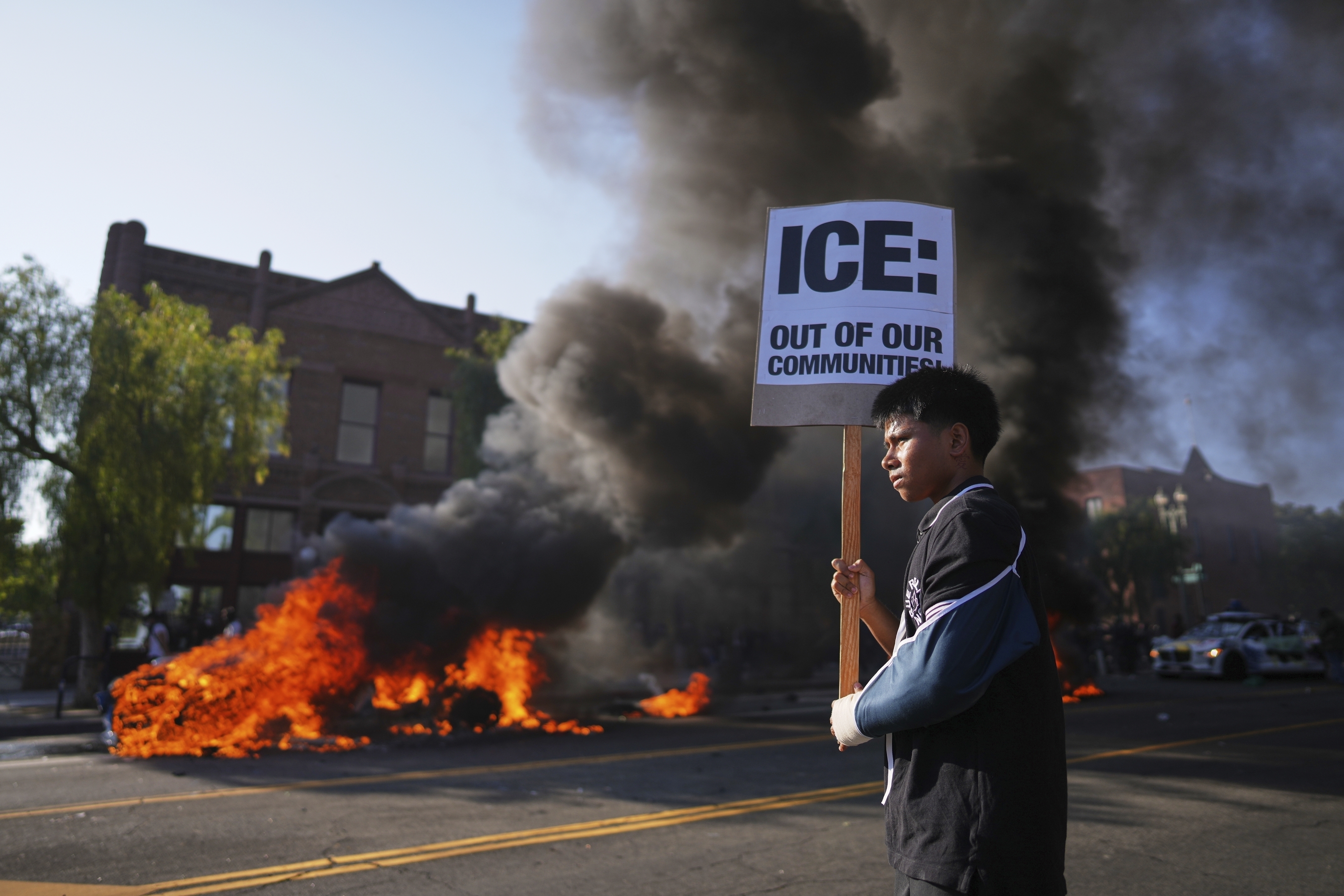 A protester holds a sign as a Waymo taxi burns near the metropolitan detention center of downtown Los Angeles, Sunday, June 8, 2025. (AP Photo/Eric Thayer)