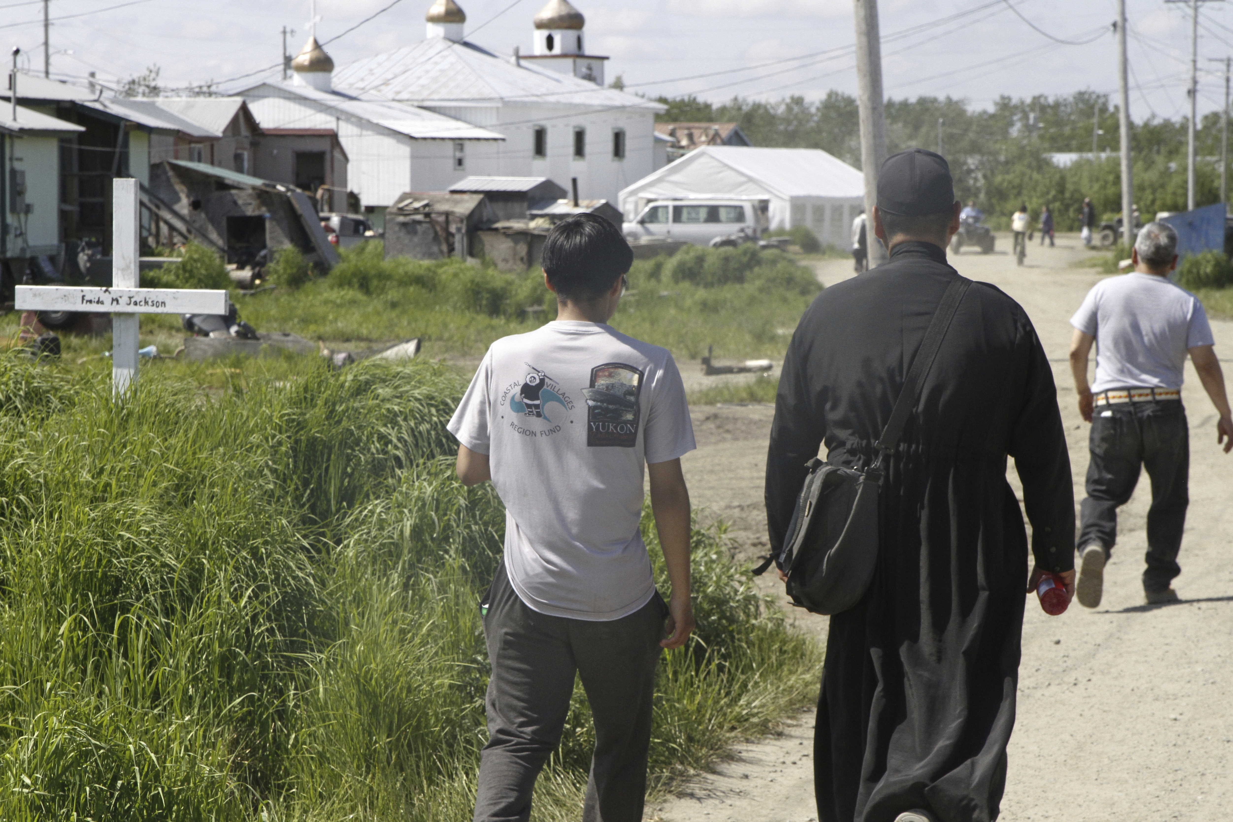 Worshippers, including an Orthodox priest wearing a black cassock, walk on the dusty streets of Kwethluk, Alaska, on June 19, 2025, heading to St. Nicholas Orthodox Church for the canonization ceremony of St. Olga, the first female Orthodox saint in North America. (AP Photo/Mark Thiessen)