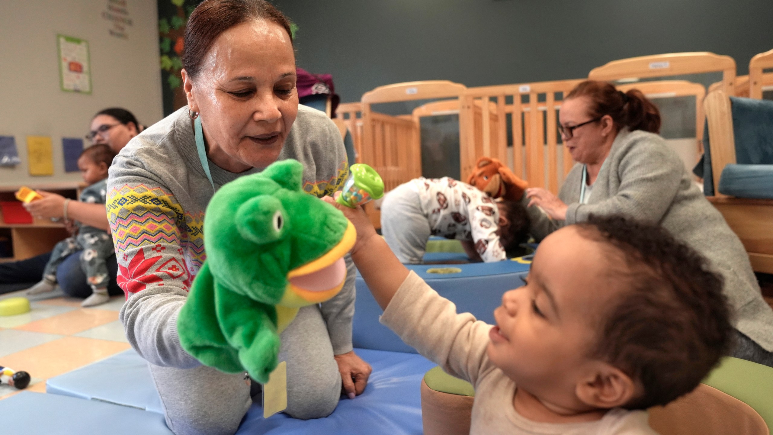 FILE - Volunteer Ydalina Luna Sosa, 62, of Providence, R.I., front left, uses a frog puppet for entertainment while supervising 10-month-old Leo Morgenweck, front right, in an early childcare program at Federal Hill House, Nov. 12, 2024, in Providence, R.I. (AP Photo/Steven Senne, File)