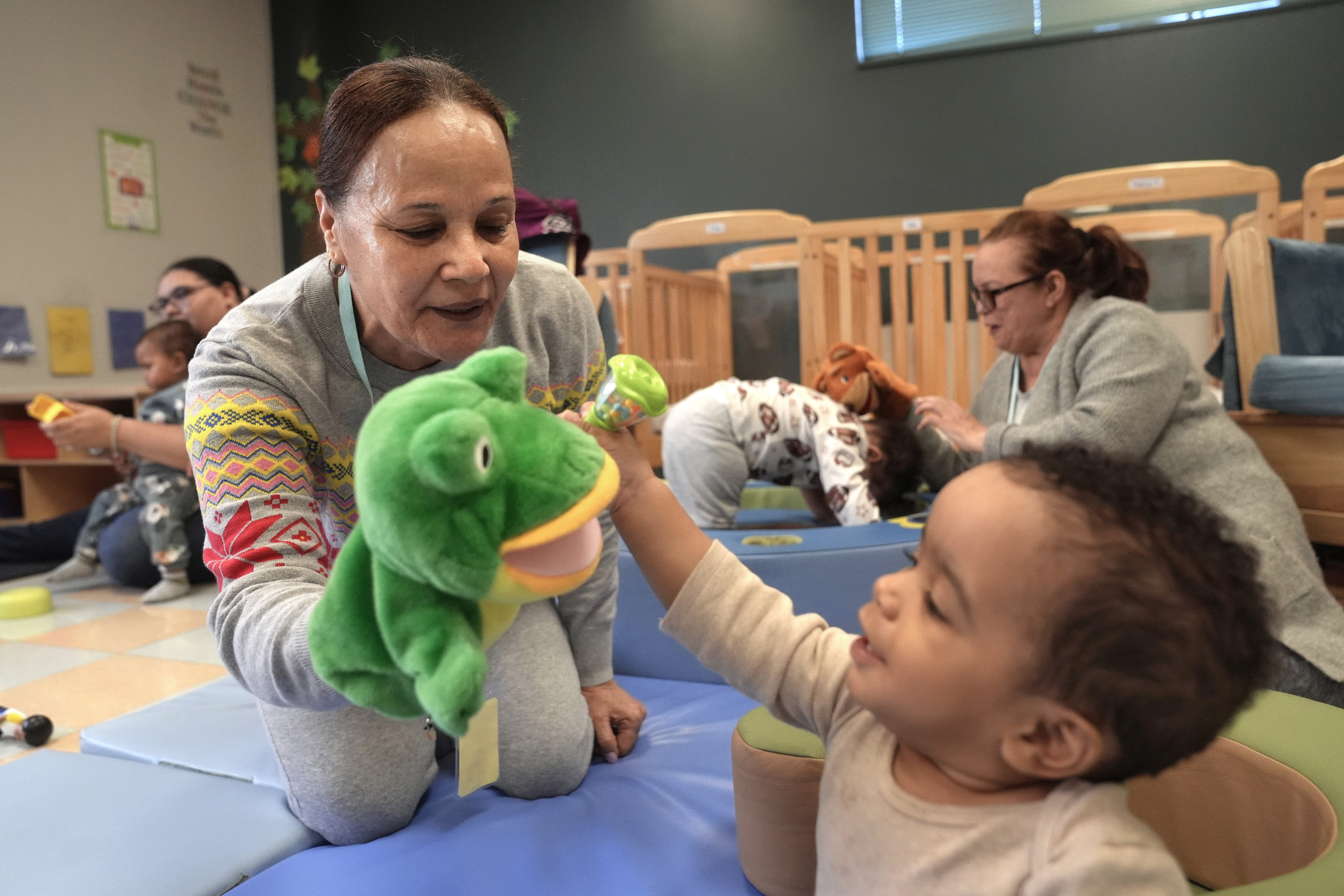 FILE - Volunteer Ydalina Luna Sosa, 62, of Providence, R.I., front left, uses a frog puppet for entertainment while supervising 10-month-old Leo Morgenweck, front right, in an early childcare program at Federal Hill House, Nov. 12, 2024, in Providence, R.I. (AP Photo/Steven Senne, File)