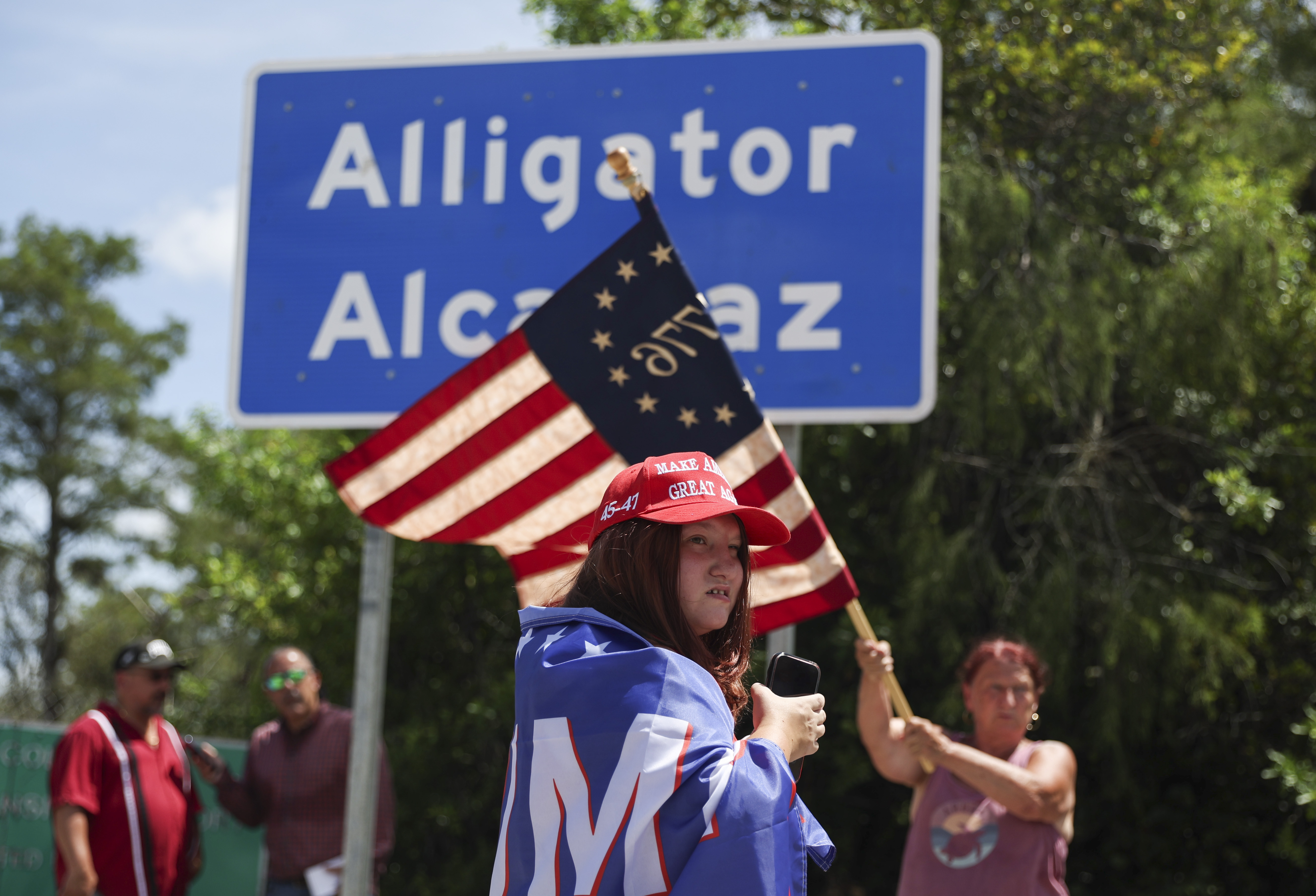 Dianne Mourer waves an American flag as Rana Mourer stands in front of a sign reading "Alligator Alcatraz" outside the Dade-Collier Training and Transition Facility, Saturday, July 12, 2025, in Ochopee, Fla. (AP Photo/Alexandra Rodriguez)