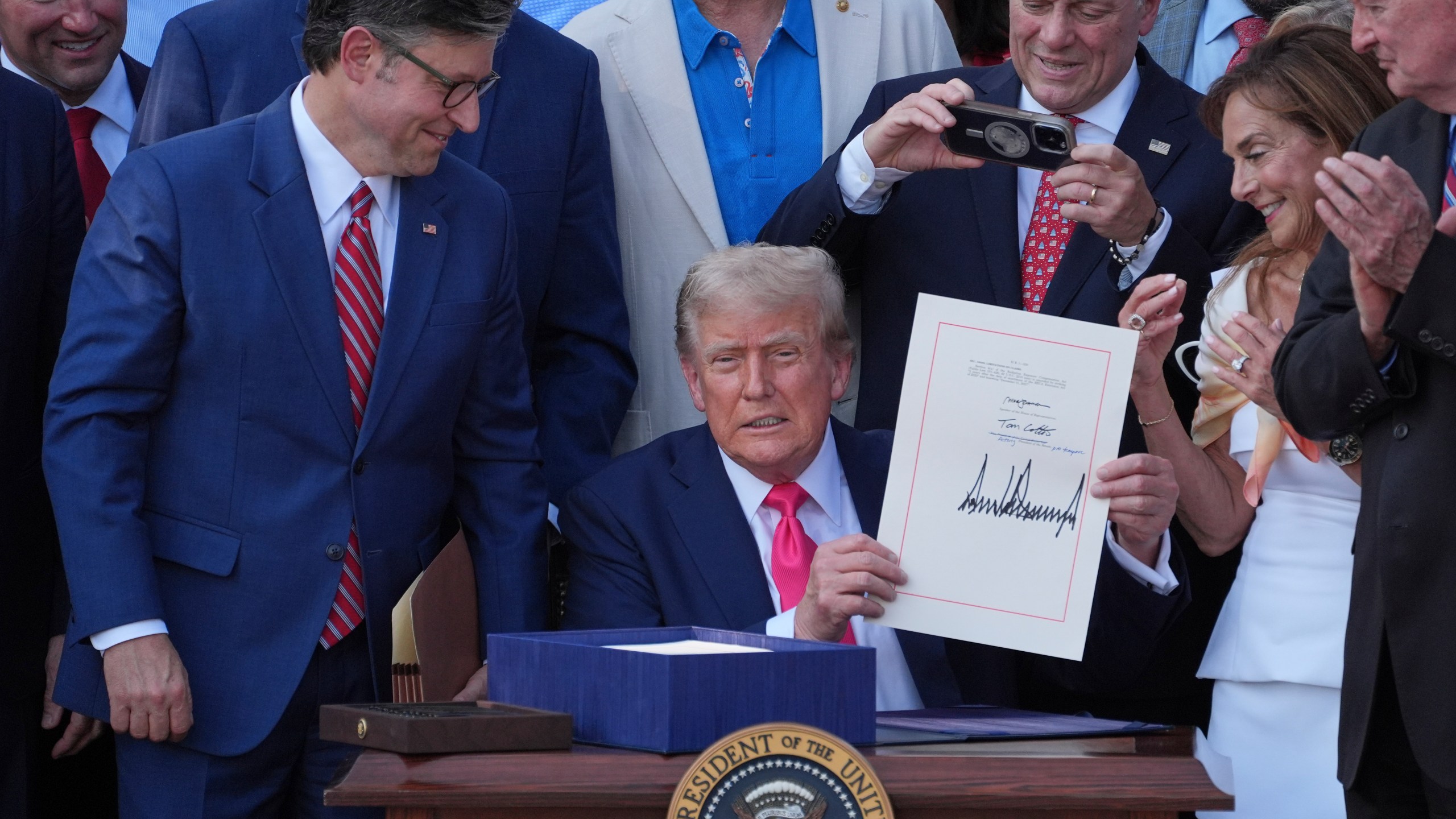 President Donald Trump holds his signed signature bill of tax breaks and spending cuts at the White House, Friday, July 4, 2025, in Washington, as House Speaker Mike Johnson of La., left, watches and Rep. Steve Scalise, R-La., takes a photo. (AP Photo/Evan Vucci)
