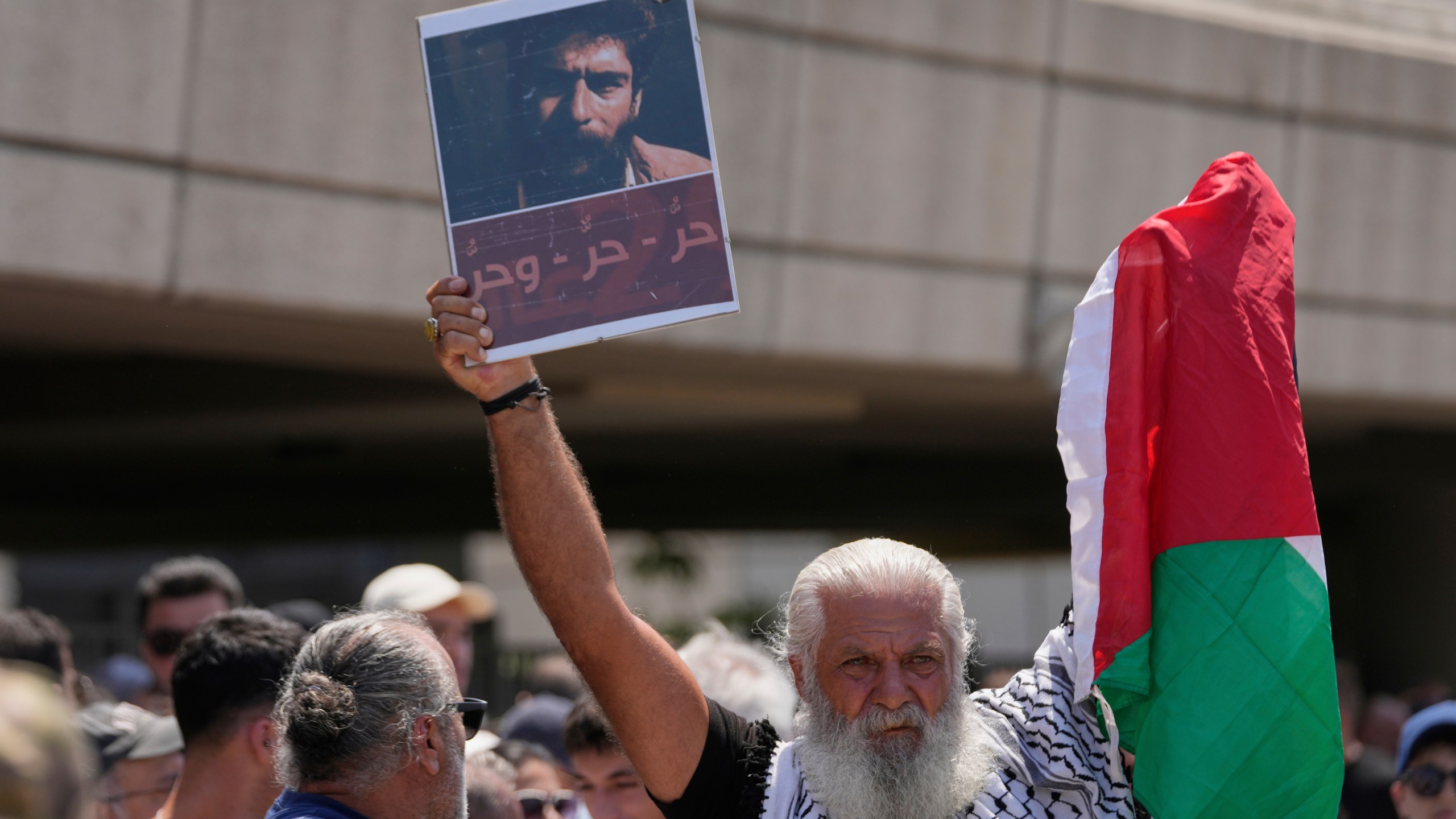 A supporter of Lebanese pro-Palestinian militant George Abdallah, who was serving a life sentence for complicity in the 1982 murders of an American and an Israeli diplomat in Paris, holds up a poster with his portrait and a Palestinian flag as he waits for Abdallah's arrival at Rafik Hariri International Airport in Beirut, Lebanon, Friday, July 25, 2025. The Arabic reads: "Free, free, and free" (AP Photo/Hussein Malla)