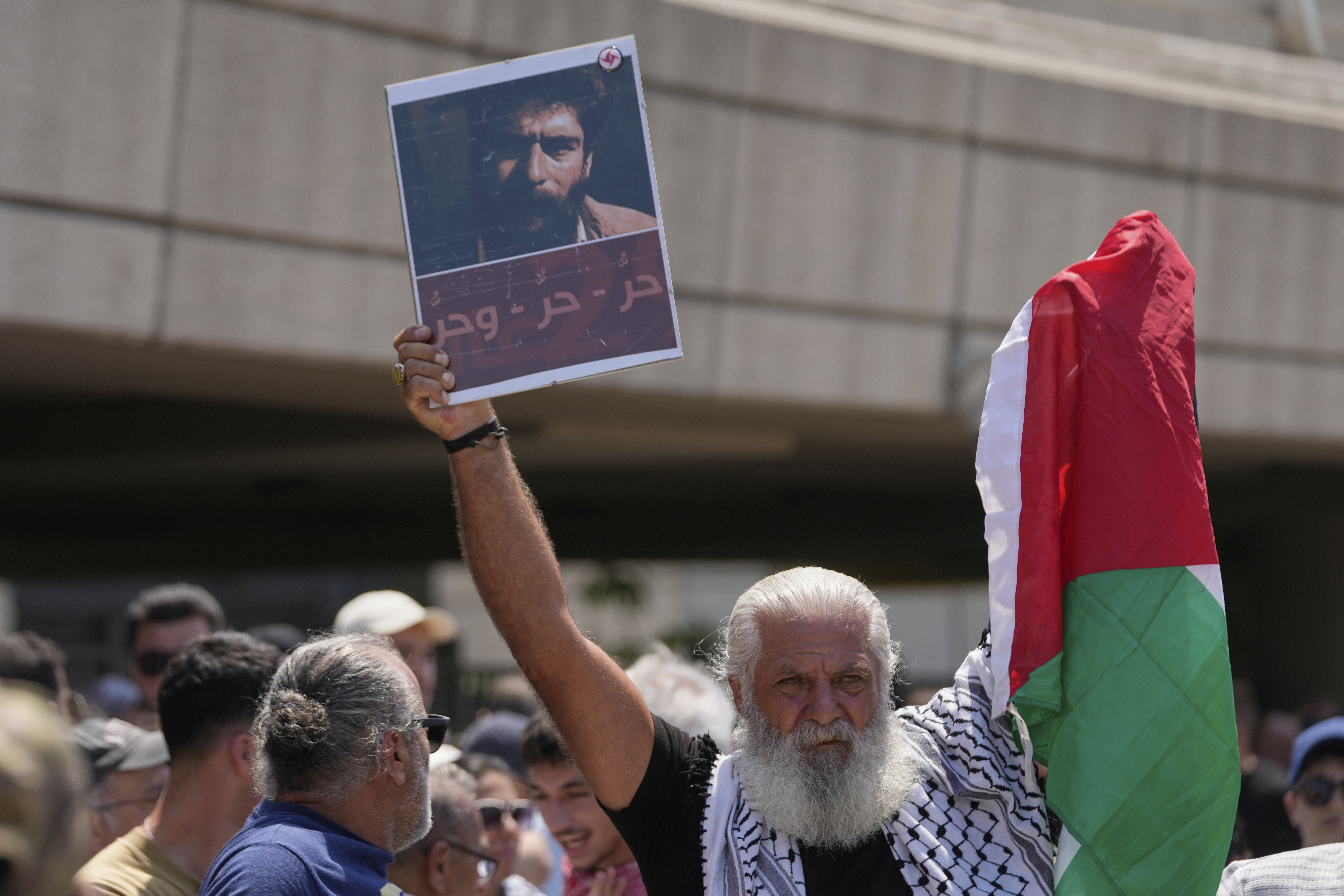 A supporter of Lebanese pro-Palestinian militant George Abdallah, who was serving a life sentence for complicity in the 1982 murders of an American and an Israeli diplomat in Paris, holds up a poster with his portrait and a Palestinian flag as he waits for Abdallah's arrival at Rafik Hariri International Airport in Beirut, Lebanon, Friday, July 25, 2025. The Arabic reads: "Free, free, and free" (AP Photo/Hussein Malla)
