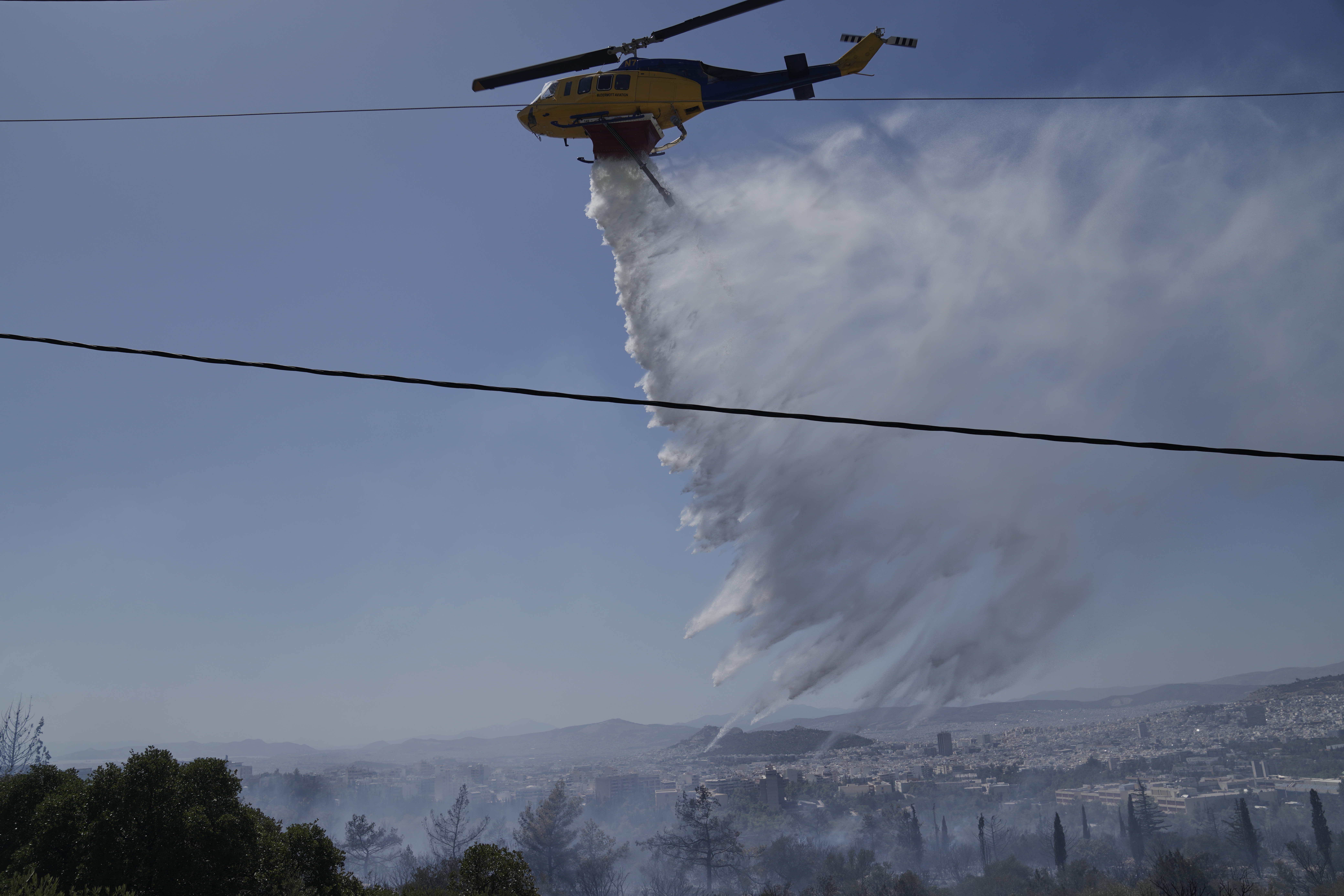 A firefighting helicopter drops water to extinguish a fire at the Polytechnic University of Athens as the Greek capital is seen in the background on Monday, July 28, 2025. (AP Photo/Thanassis Stavrakis)