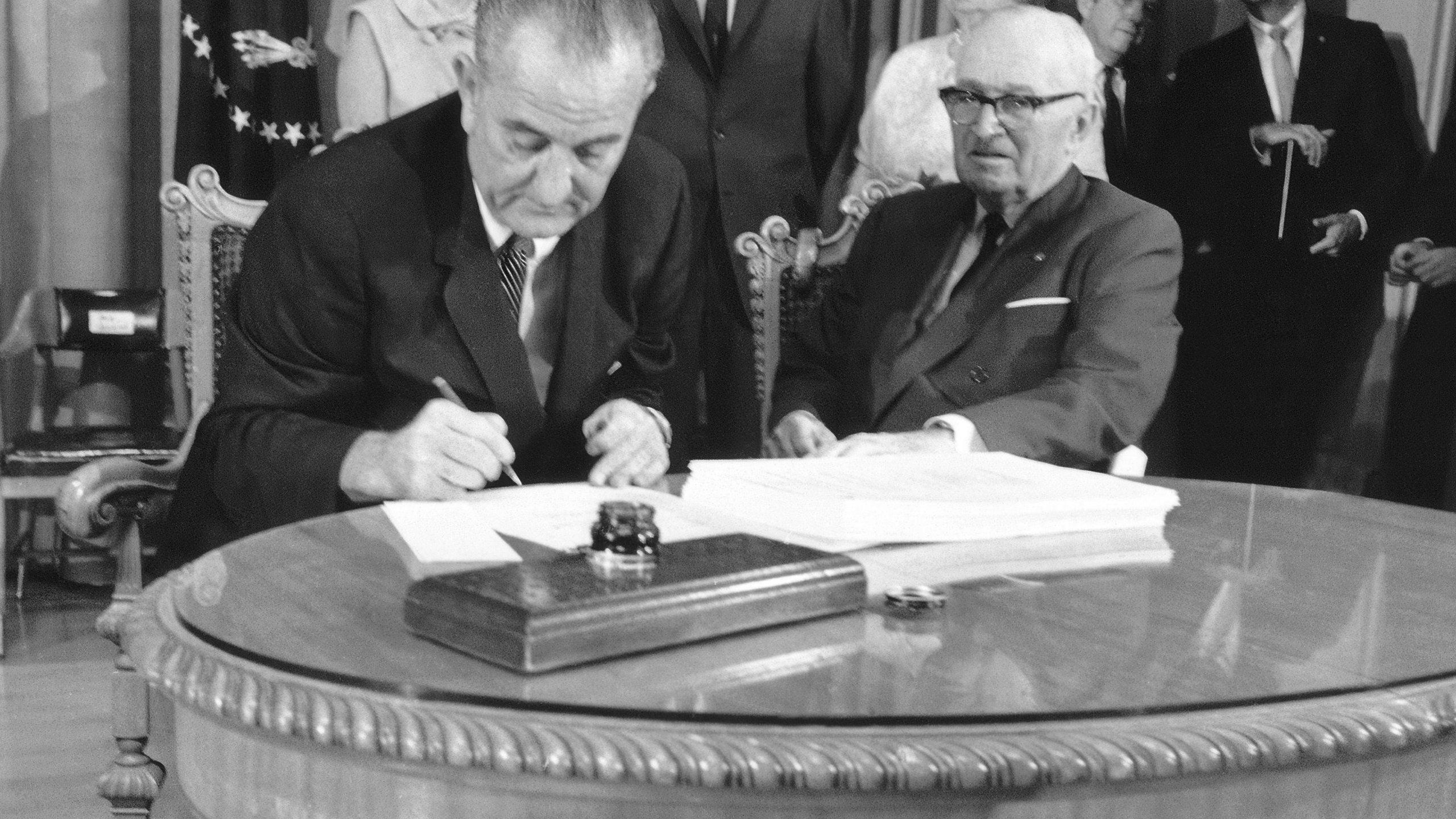 FILE - President Lyndon B. Johnson uses the last of many pens to complete the signing of the Medicare Bill into law at the Truman Library in Independence, Missouri, July 30, 1965, with former President Harry S. Truman at his side. At rear are Lady Bird Johnson, Vice President Hubert Humphrey, and former first lady Bess Truman. (AP Photo, File)