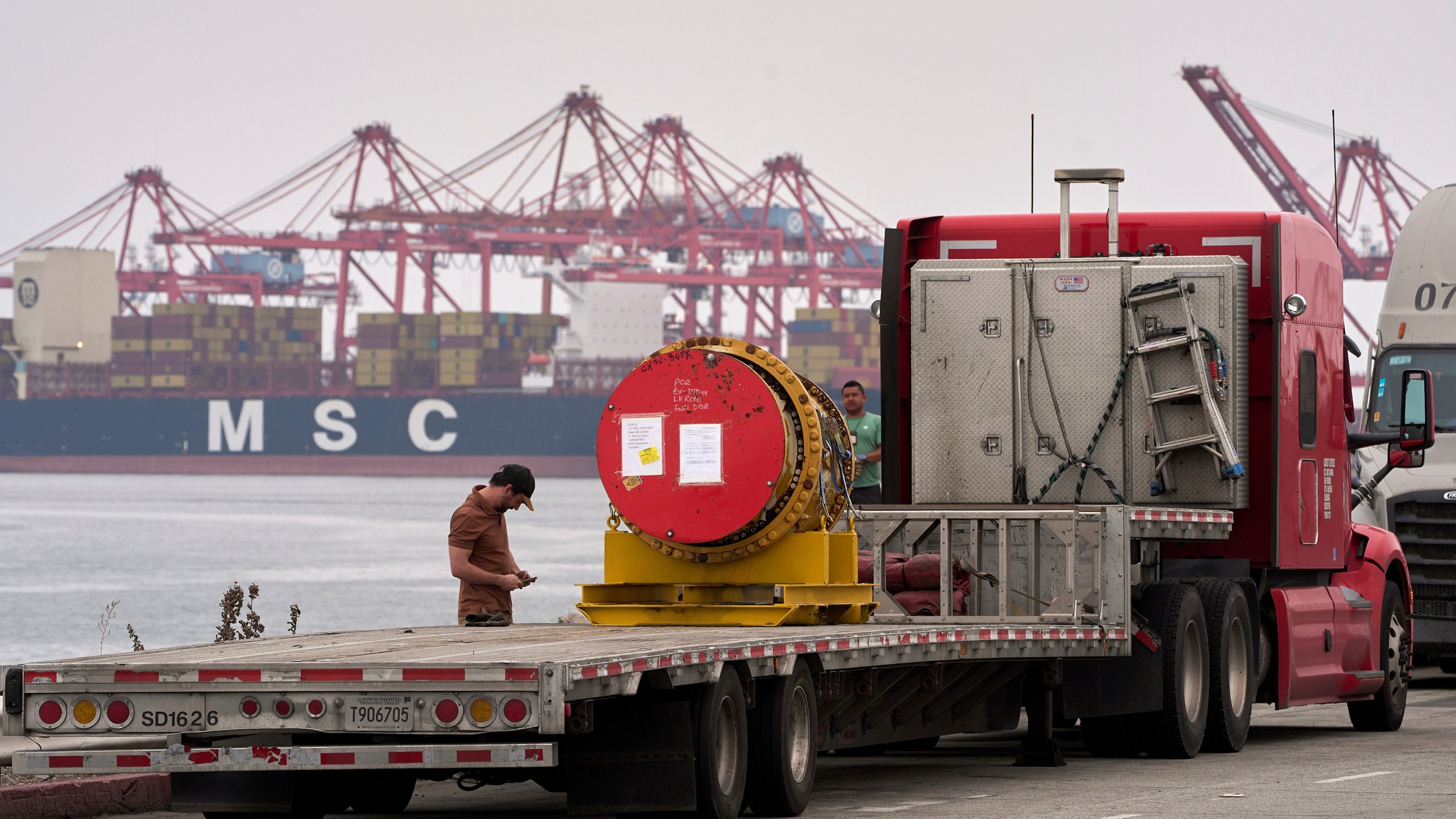 A truck driver secures a heavy machinery prior to transporting it at the Port of Long Beach, Calif., Friday, Aug. 1, 2025. (AP Photo/Damian Dovarganes)