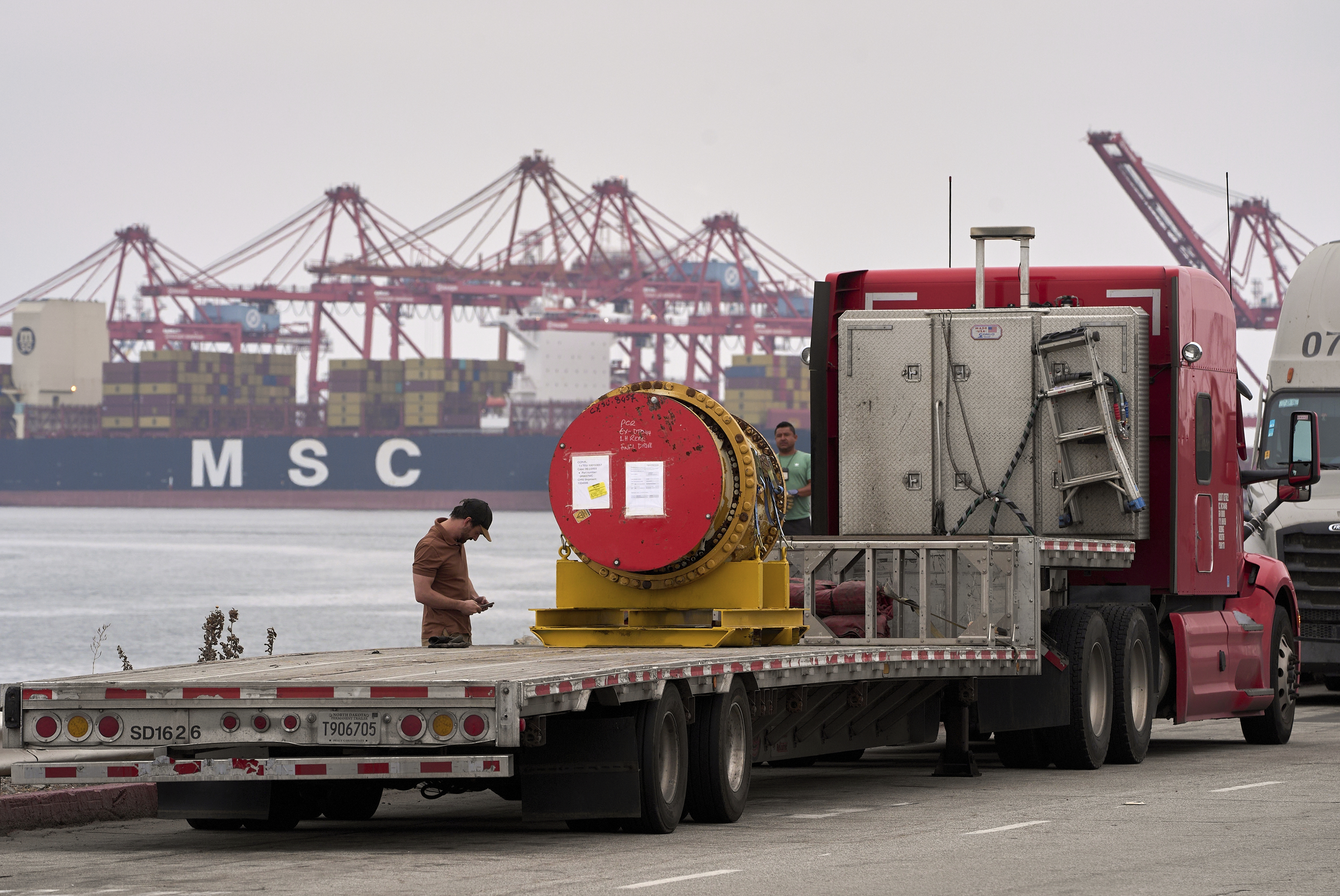 A truck driver secures a heavy machinery prior to transporting it at the Port of Long Beach, Calif., Friday, Aug. 1, 2025. (AP Photo/Damian Dovarganes)