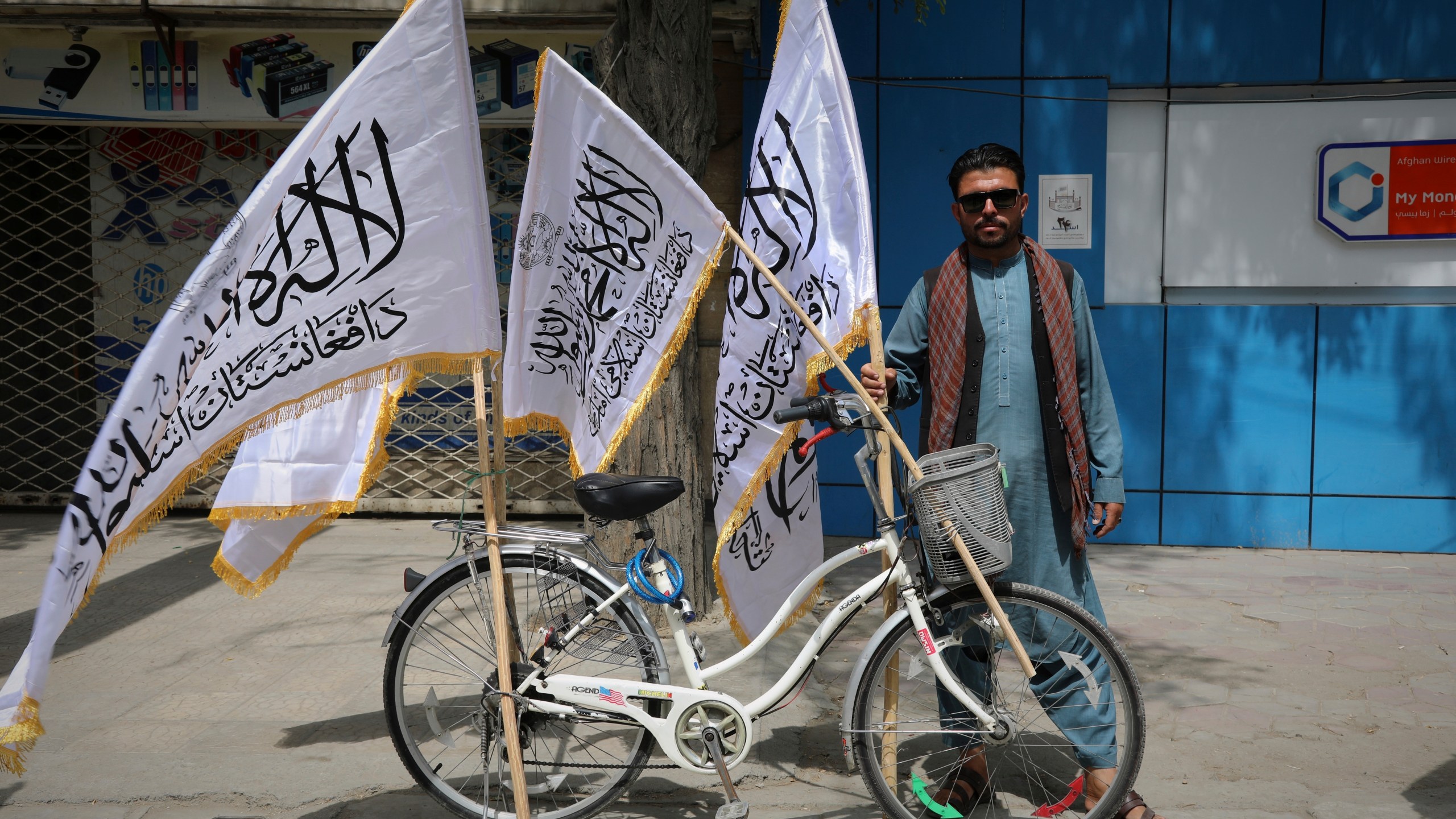 A man poses for the camera next to his bicycle while selling Taliban flags ahead of celebrations marking the fourth anniversary of the U.S. withdrawal from Afghanistan and the beginning of Taliban rule in Kabul, Afghanistan, Thursday, Aug. 14, 2025. (AP Photo/Siddiqullah Alizai)
