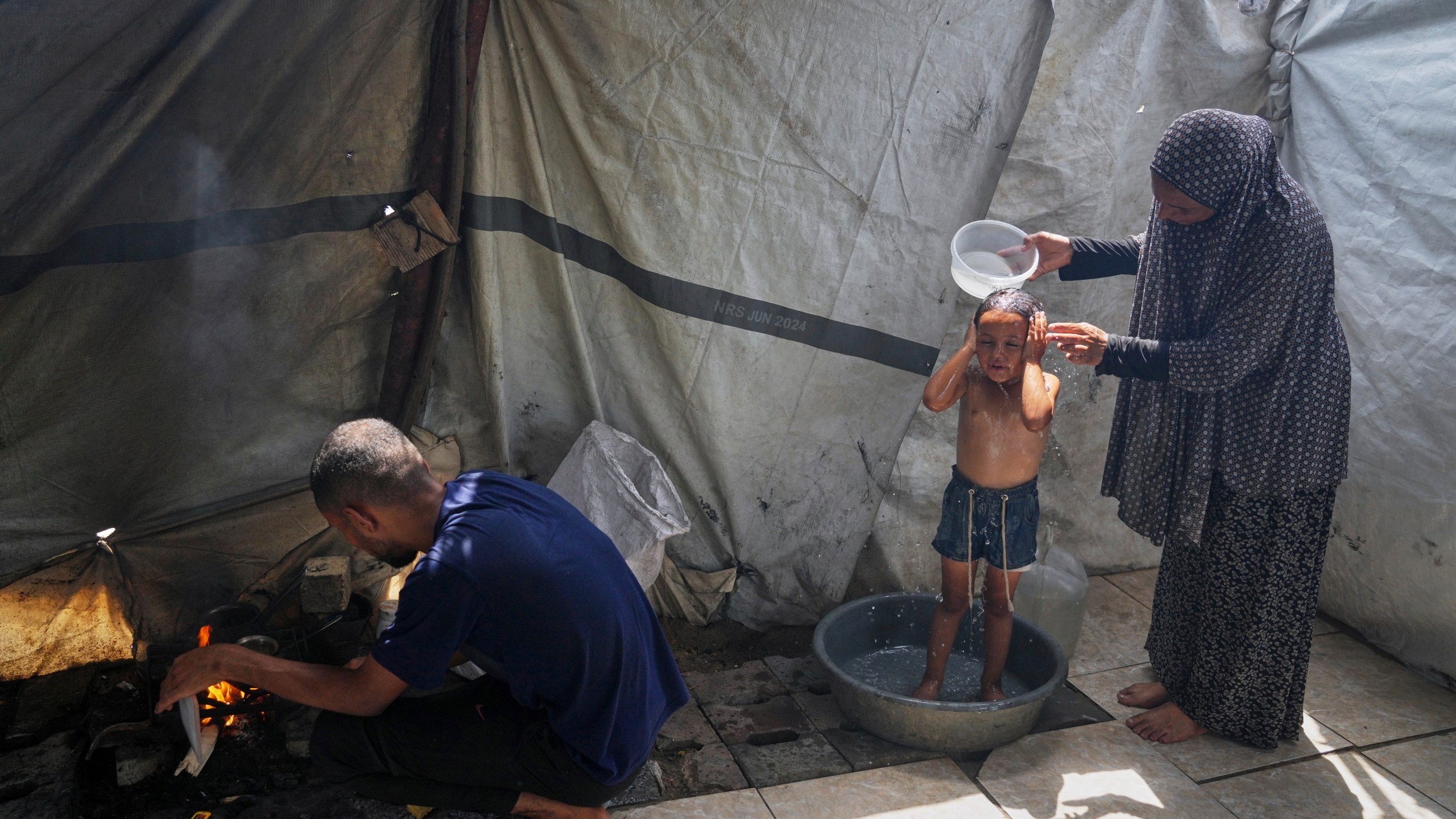 Enaam Al Majdoub uses water collected from a distribution point to bathe her 3-year-old granddaughter, Jourieh, while her son Zaki uses some of the water for cooking in their family tent in Gaza City on Tuesday, August 12, 2025. (AP Photo/Jehad Alshrafi)