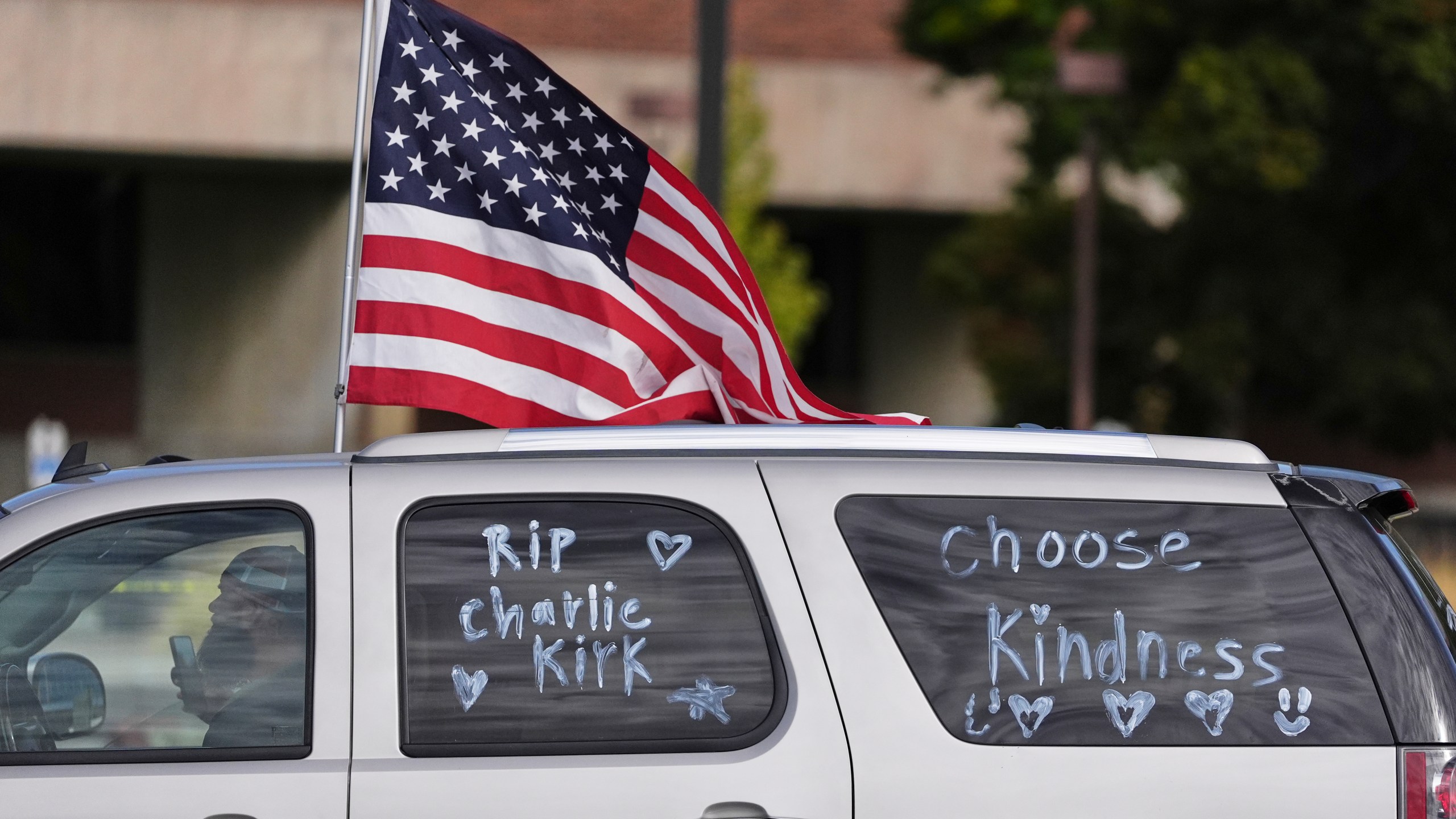 A vehicle marked with messages written on its windows in tribute to Turning Point USA CEO and co-founder Charlie Kirk and carrying an American flag, drive past at Utah Valley University, Saturday, Sept. 13, 2025, in Orem, Utah. (AP Photo/Lindsey Wasson)