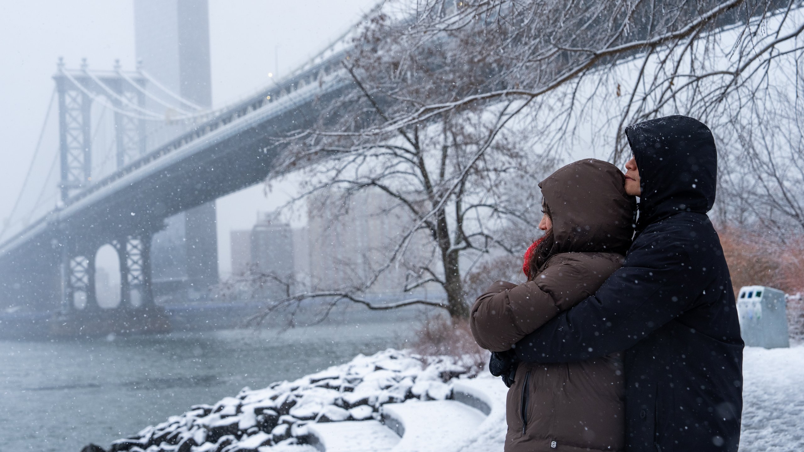 A couple embrace during a snow in front of the Manhattan bridge, Sunday, Dec. 14, 2025, in the Brooklyn Borough of New York. (AP Photo/Adam Gray)
