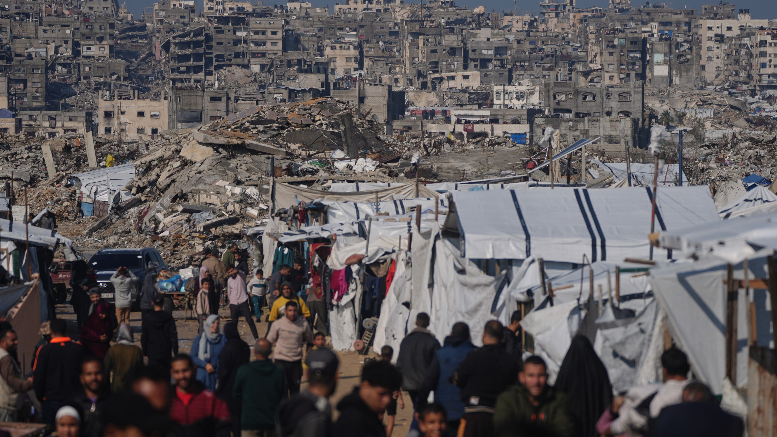 Palestinians walk along a street past a tent camp, backdropped by buildings destroyed during Israeli air and ground operations in Gaza City, Wednesday, Dec. 17, 2025. (AP Photo/Jehad Alshrafi)